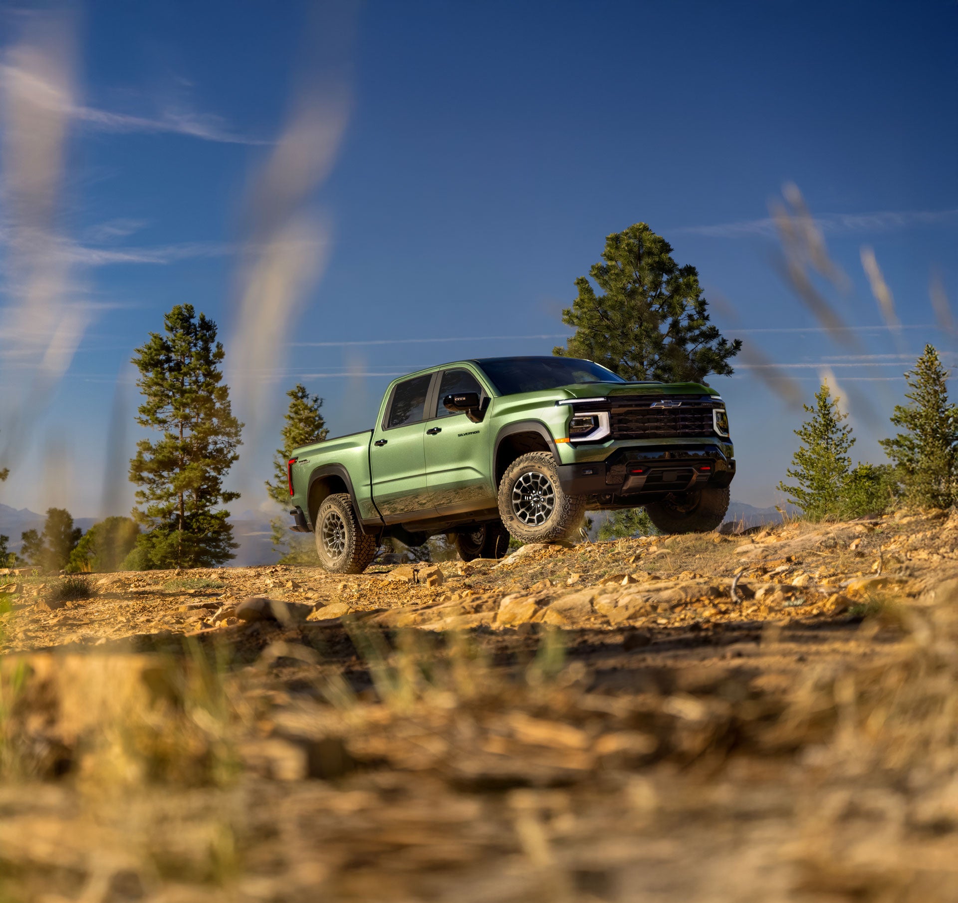 A gray 2027 Chevrolet Silverado 1500 with distinctive orange daytime running lights parked on a dirt road in a vast, arid landscape under a bright sky.